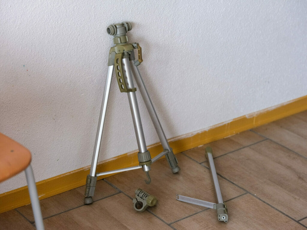 Ein Kamerastativ auf dem Boden neben einem Stuhl hat offensichtlich Schäden davongetragen: einer seiner Streben ist abgebrochen.
