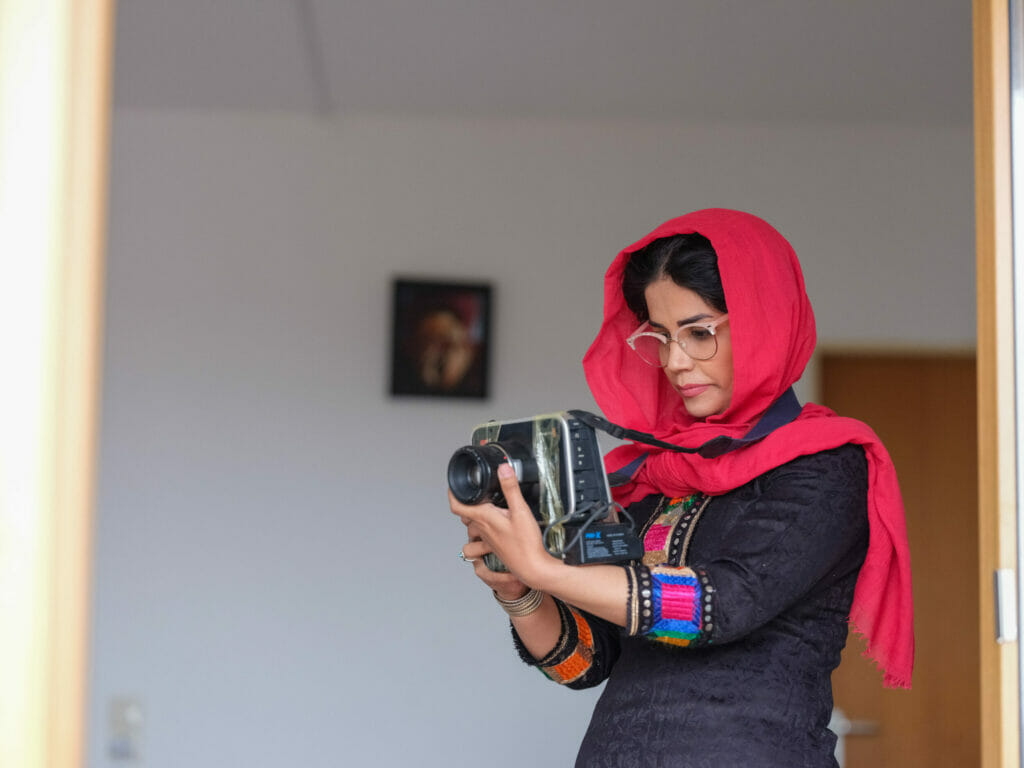 Eine junge Frau trägt ein rotes Kopftuch, unter dem dunkle lockige Haare hervorlugen, dazu ein traditionell afghanisches Gewand mit schwarzer Grundfarbe und rot-pink-violetten Verzierungen an den Ärmeln. Sie ist im Profil zu sehen und schaut konzentriert auf die Kamera, die sie vor sich hält. Im Hintergrund hängt ein kleines Bild an der Wand, das nur unscharf zu sehen ist. Zainab Entezar zählt zu den ersten Afghan:innen, die über das Bundesaufnahmeprogramm nach Deutschland evakuiert wurden.