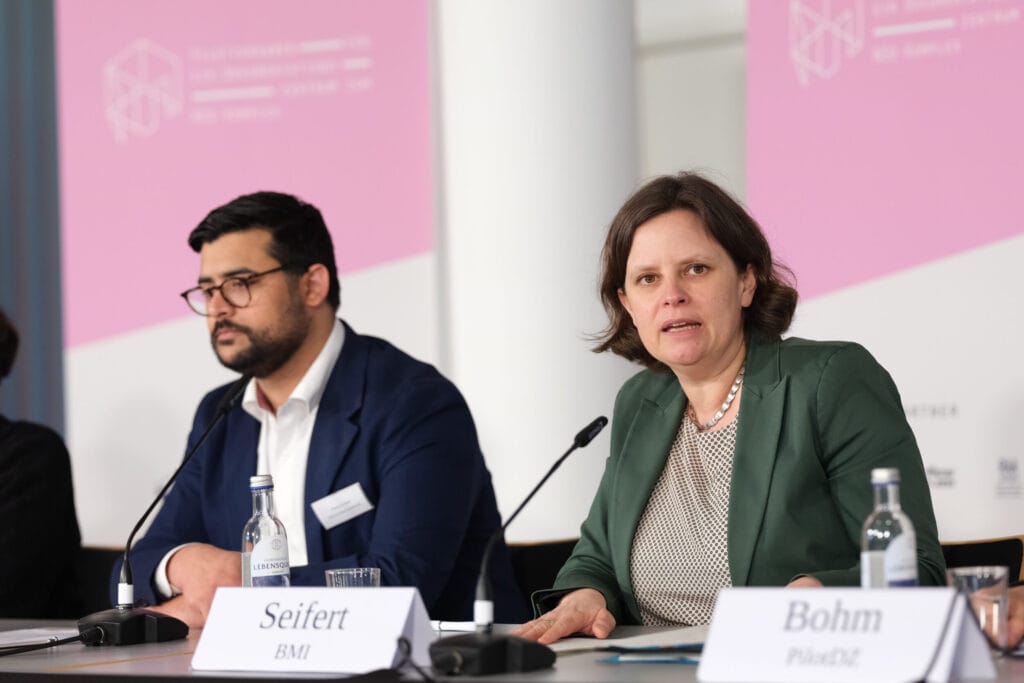 Two panelists, a woman and a man, discuss during a conference, with name tags and microphones in front of them.