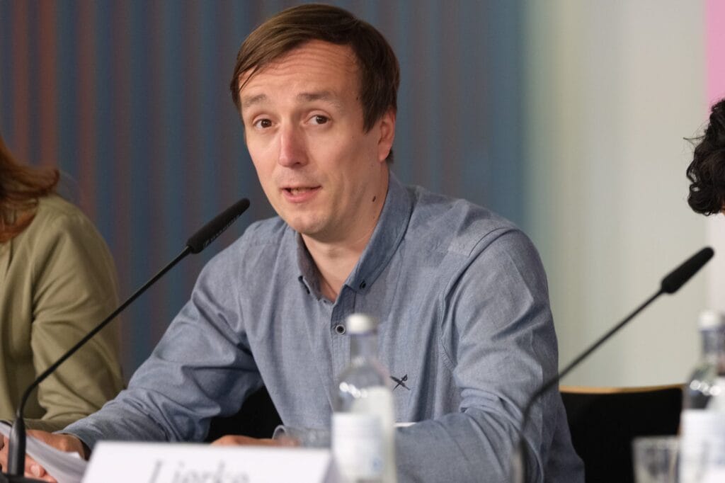 A man in a blue shirt speaks at a conference. In front of him stands a table with microphones and a name badge.