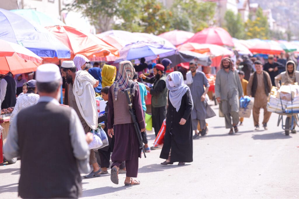 Ein überfüllter Markt im Freien in Kabul, Afghanistan, mit bunten Regenschirmen und Passanten. Eine Person trägt eine Waffe, eine andere schiebt einen mit Kisten beladenen Karren.