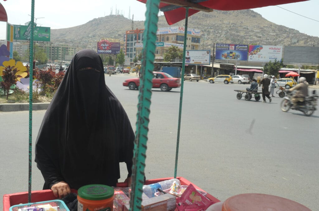 Eine Frau in einem schwarzen Niqab steht an einer belebten Kreuzung in der Stadt an einem mit Waren gefüllten Straßenkarren, im Hintergrund sind Hügel und Gebäude zu sehen.